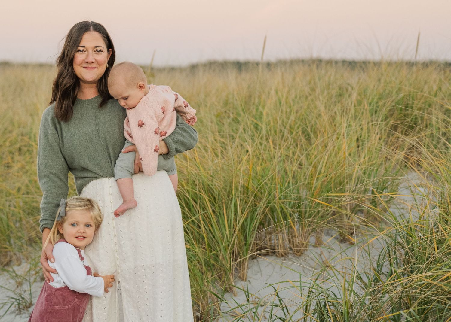 Family wearing coordinated outfits stands together in a grassy field during golden hour sunset.