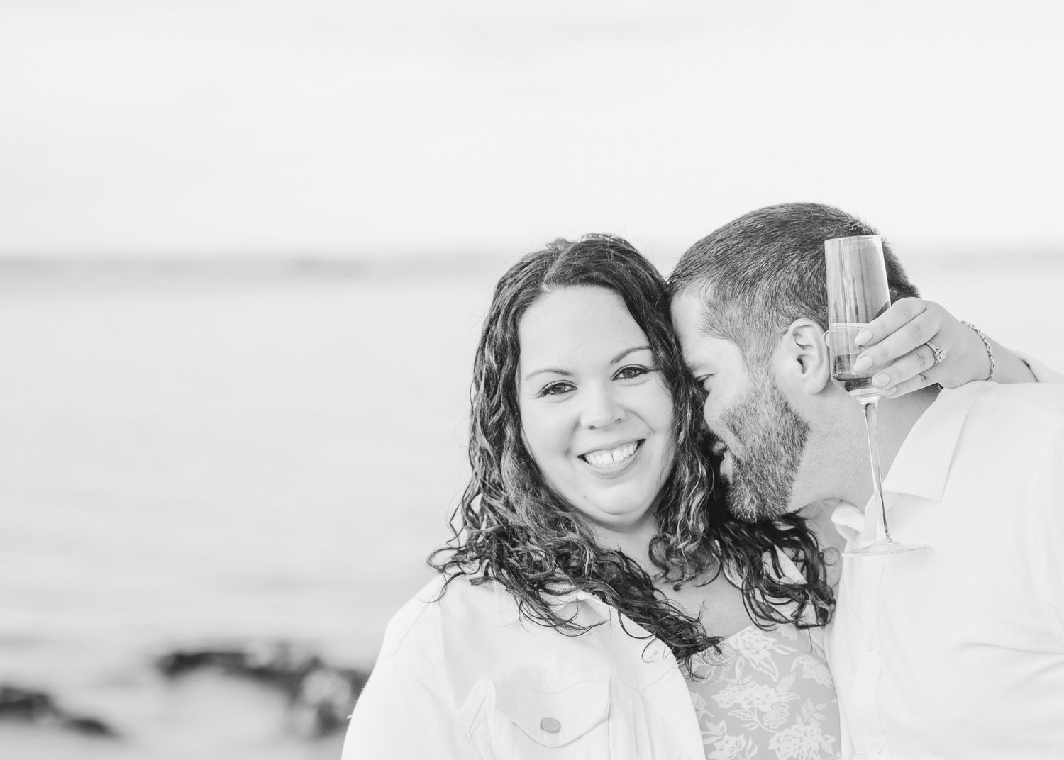 A black and white romantic portrait of a couple laughing by the ocean.