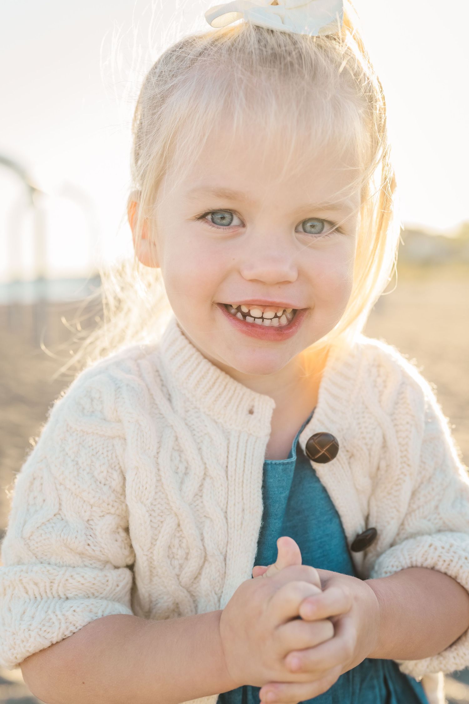 Young child wearing a cream knit cardigan smiles brightly in warm sunlight.