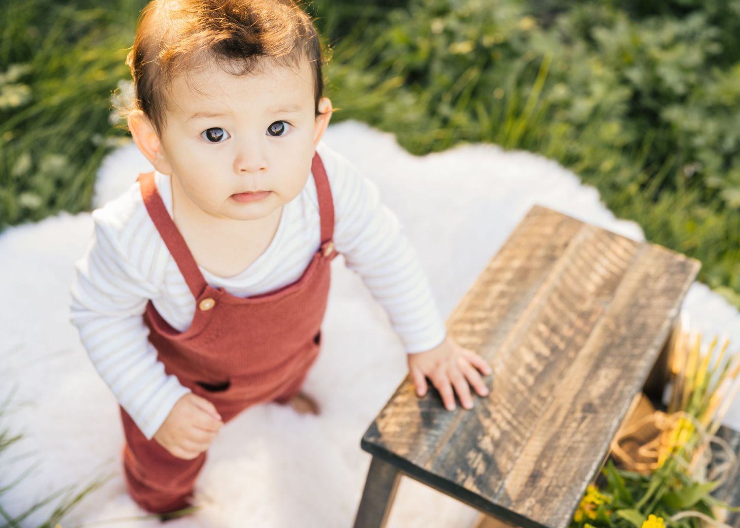 A baby in red overalls sits on a white blanket next to a wooden crate in an outdoor setting.