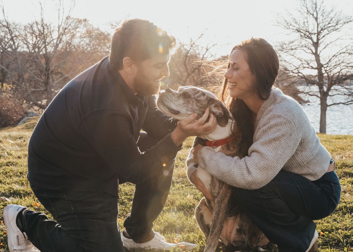 A couple outdoors sharing a moment with their brown dog in warm autumn sunlight next to trees.