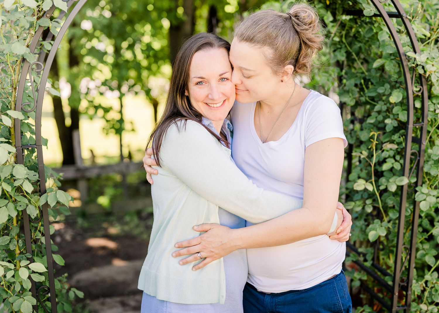 Two people share an intimate embrace under a garden archway surrounded by greenery on a sunny day.