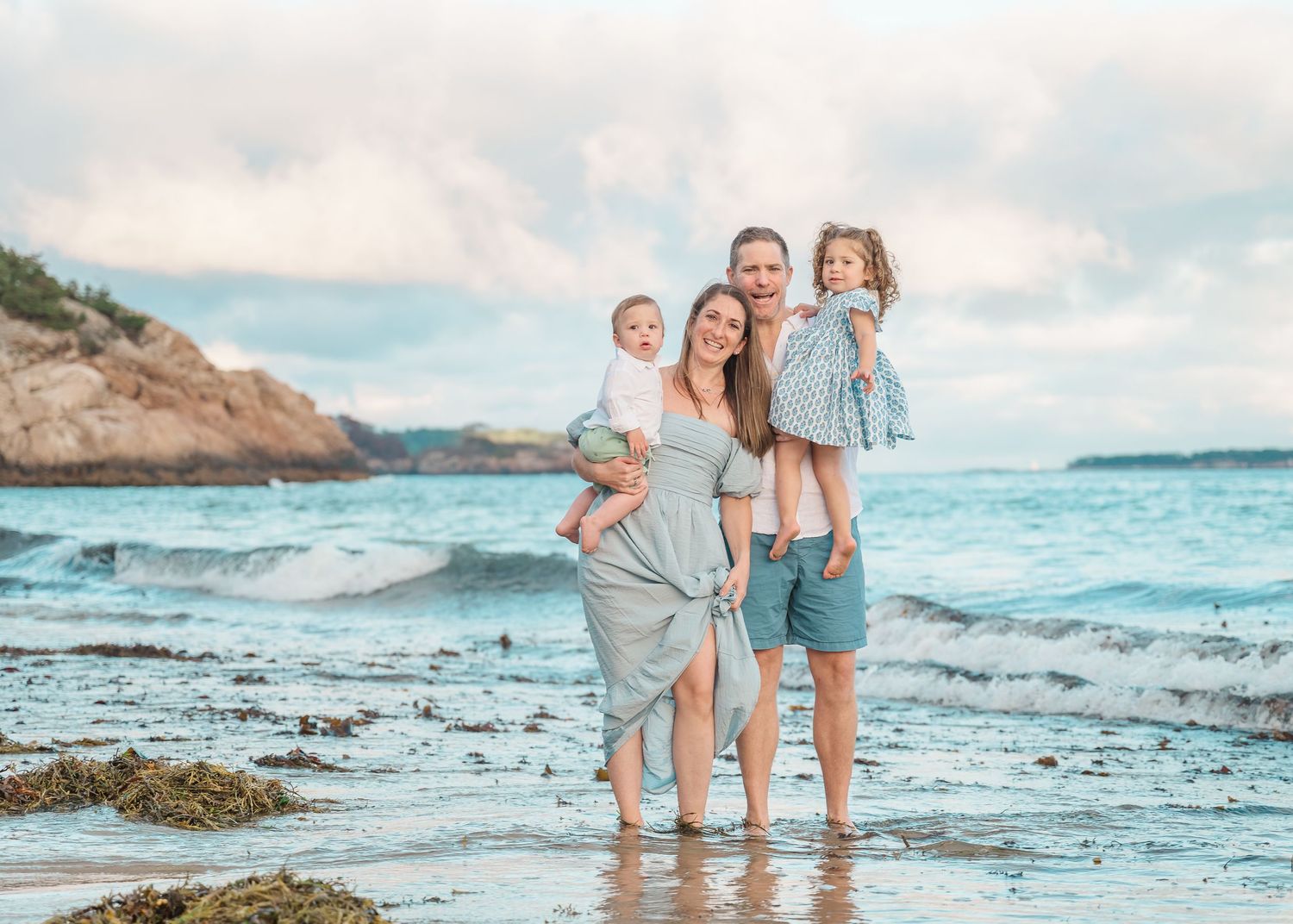 A family enjoying time together on a rocky beach with ocean waves and cloudy skies in the background.