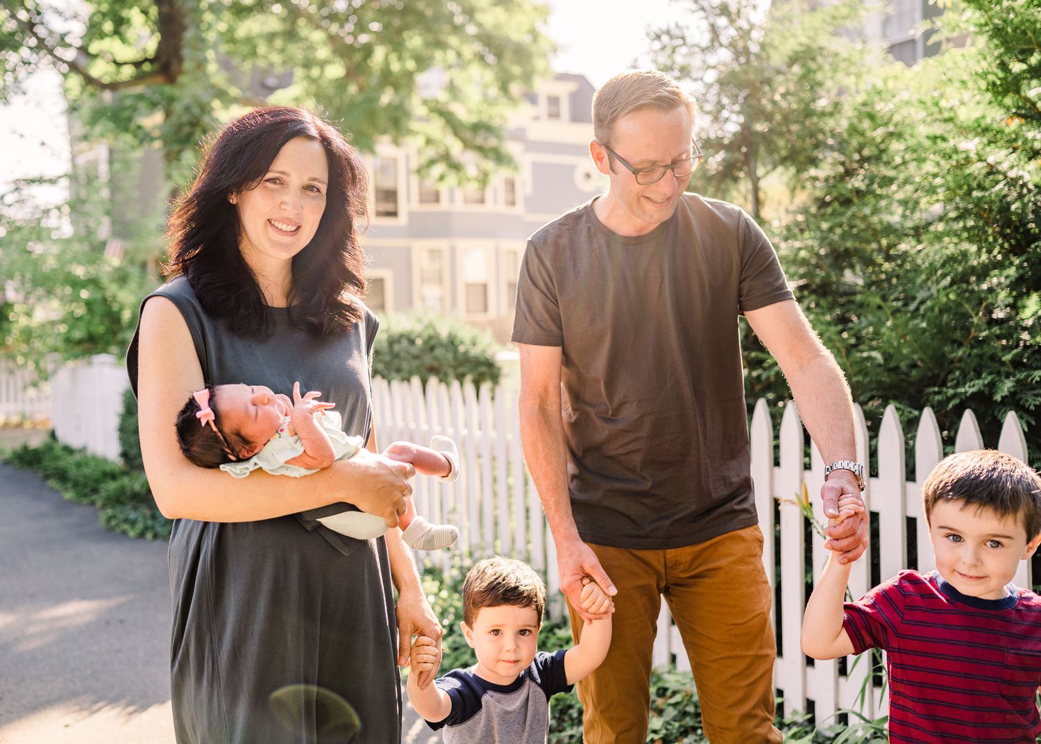 Family walking along suburban pathway with white picket fence and trees.