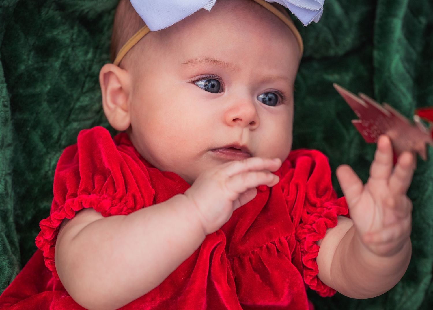 Baby in red dress and white bow reaches toward ornament against green background.