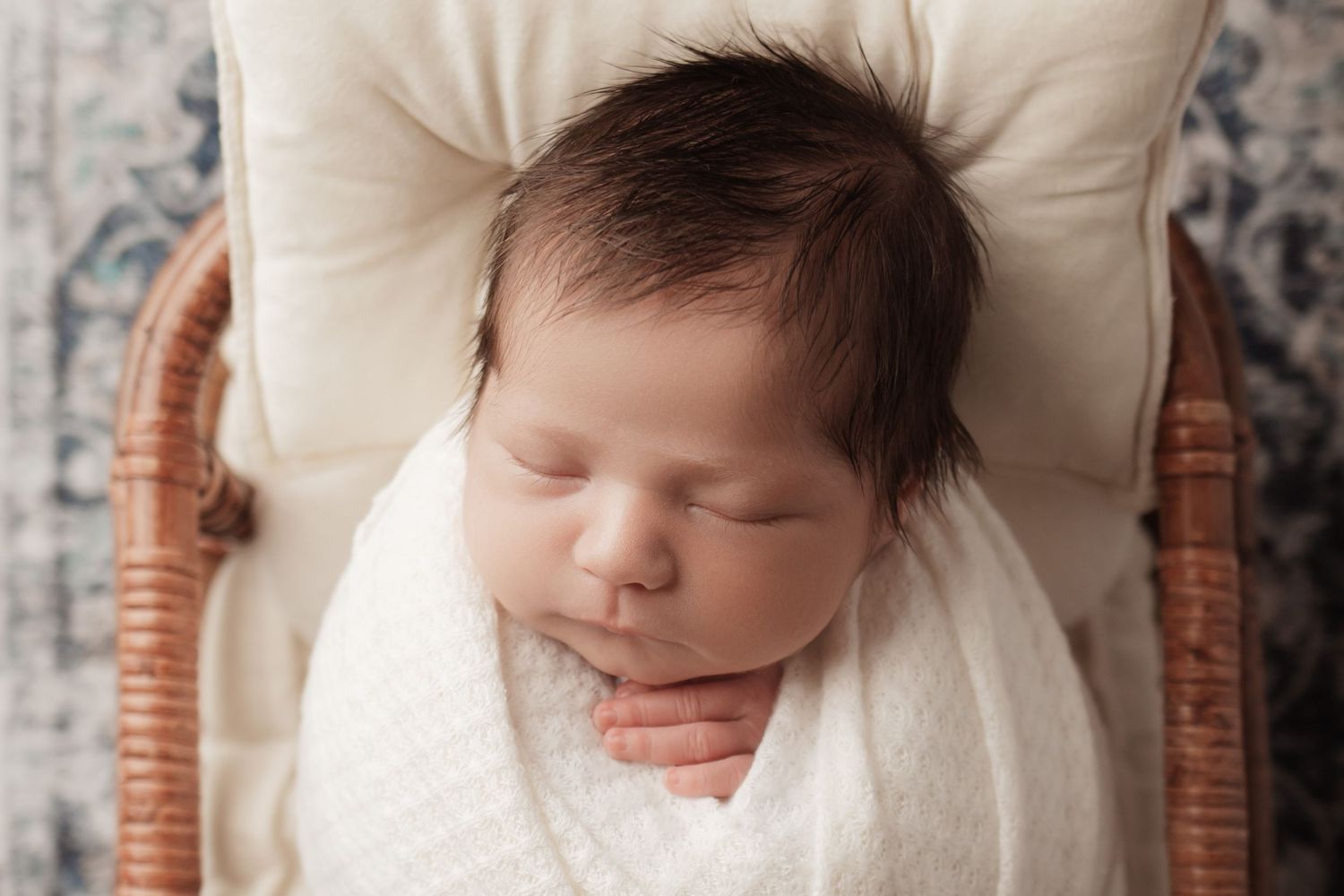 Peaceful sleeping newborn with dark hair wrapped in white swaddle in a wicker chair.
