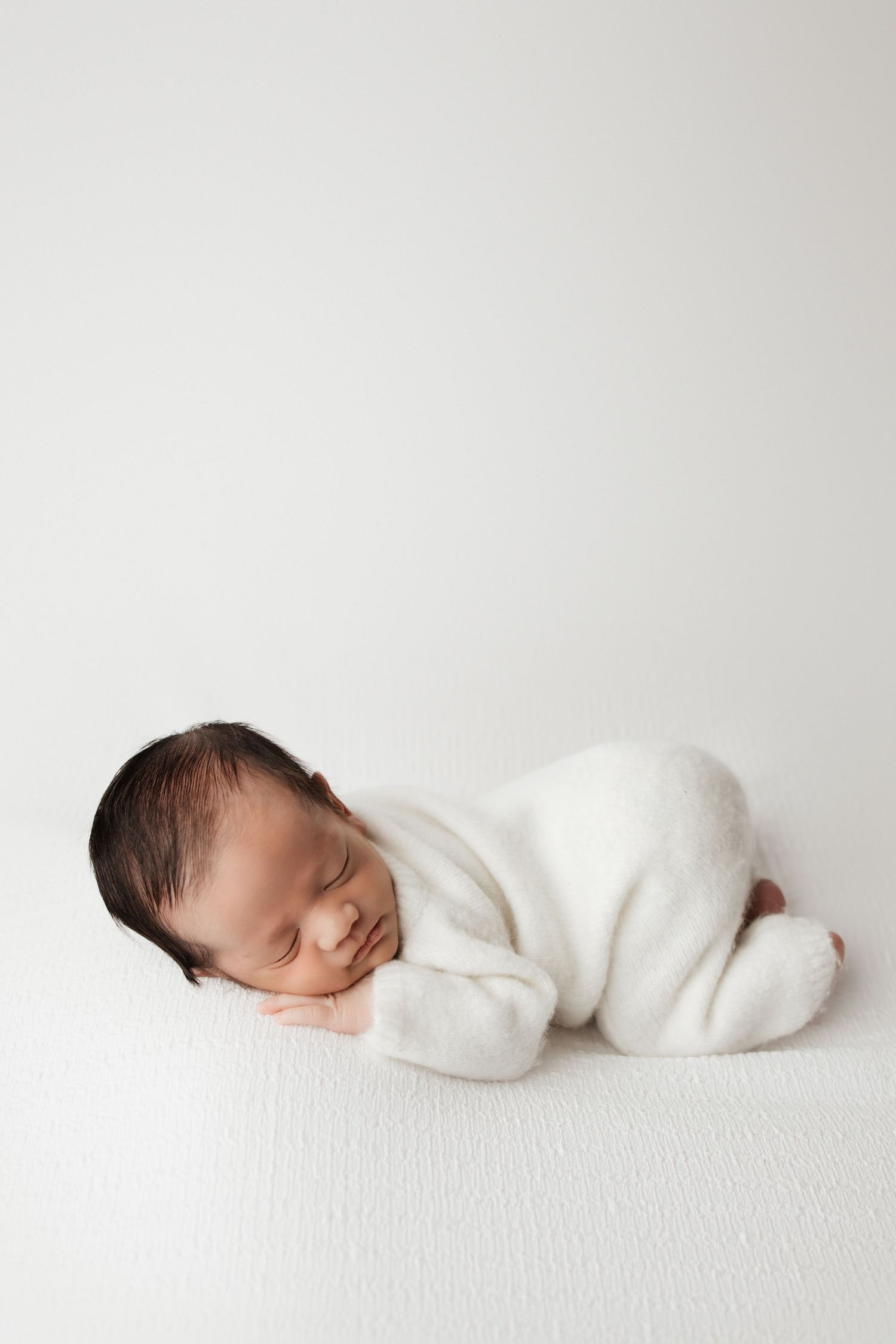 Peaceful newborn baby in white romper lying on side on white surface with minimalist background.