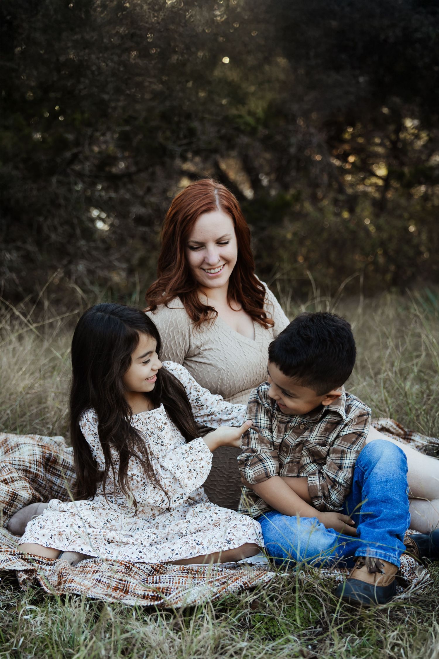 Redheaded mother sitting on blanket with two children in tall grass field.