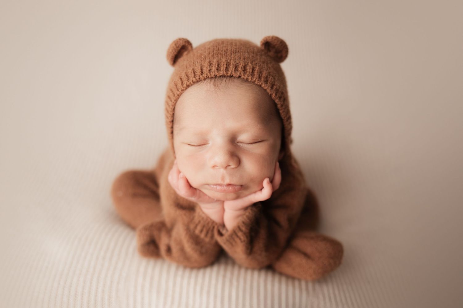 Sleeping newborn in brown knit bear outfit and bonnet posed with hands under chin.