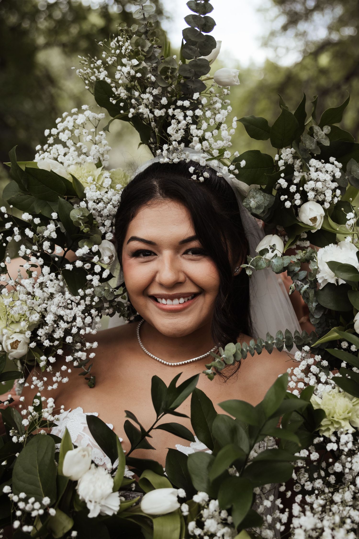 Smiling bride surrounded by white flowers and greenery, wearing a pearl necklace and veil on her wedding day.