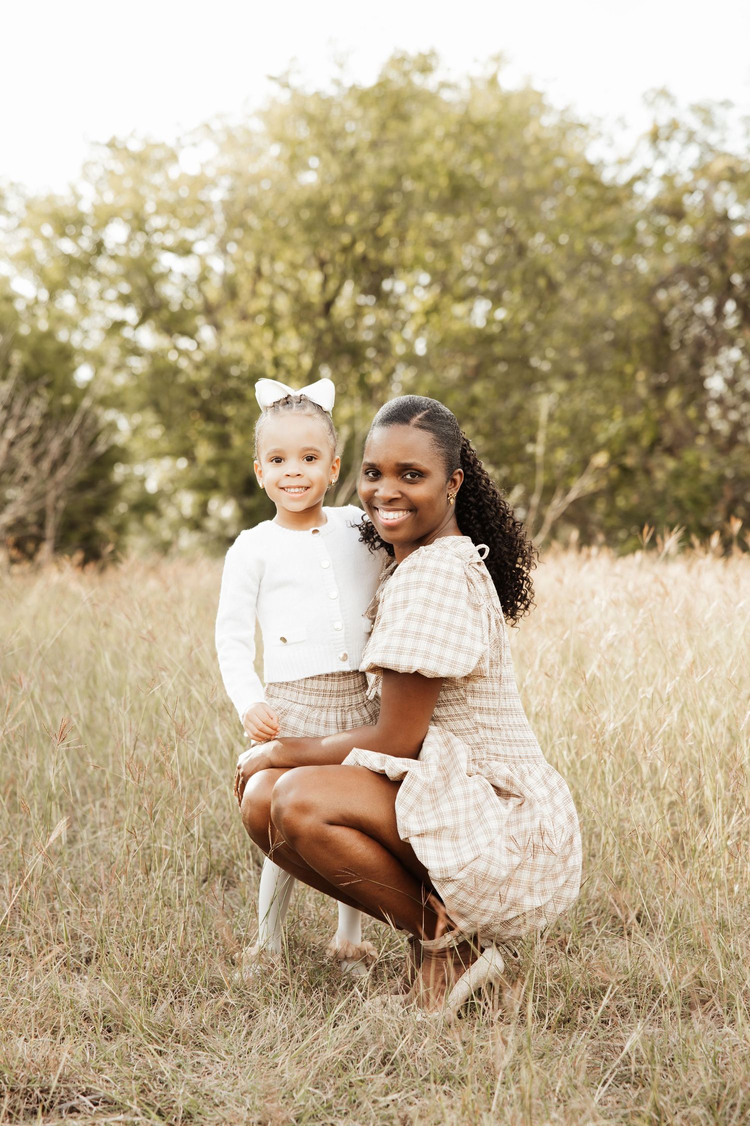 A smiling Black mother in a gingham dress crouches beside her young daughter in a field.