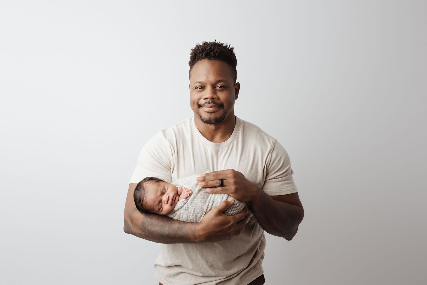 A proud father smiles warmly while cradling his swaddled newborn baby in a bright white studio setting.