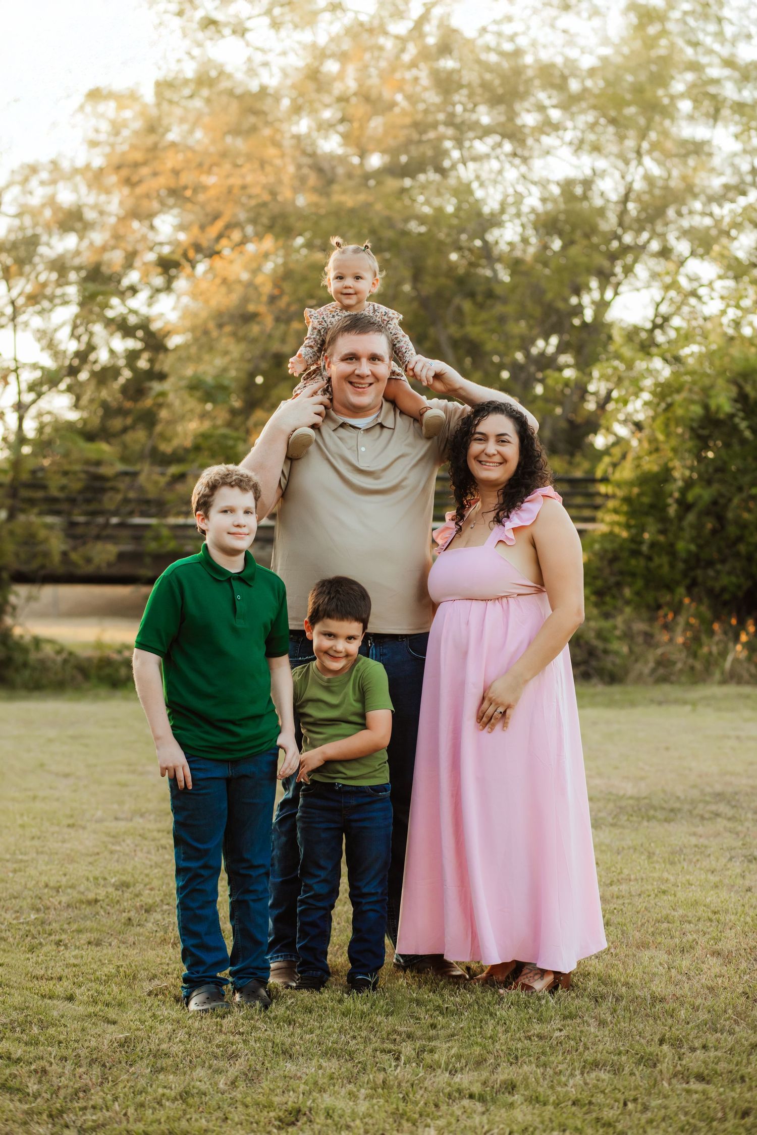 A smiling family of five poses outdoors on green grass with trees in the background at golden hour.