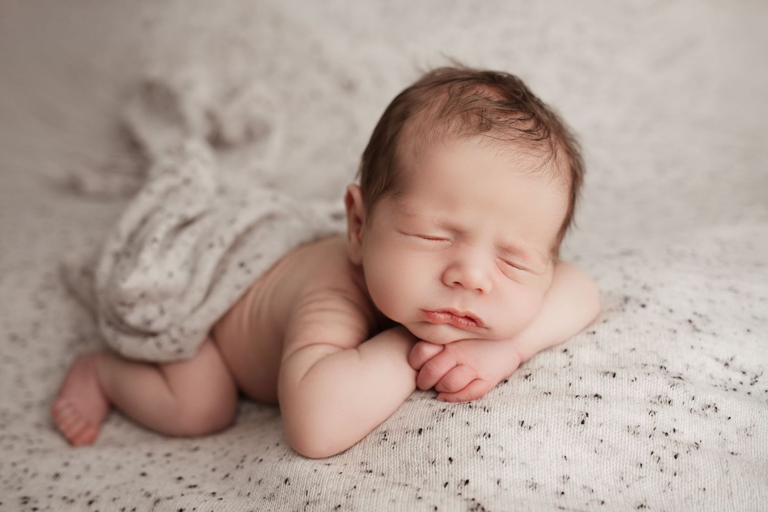 Peaceful sleeping newborn baby posed with hands under chin on white speckled blanket.