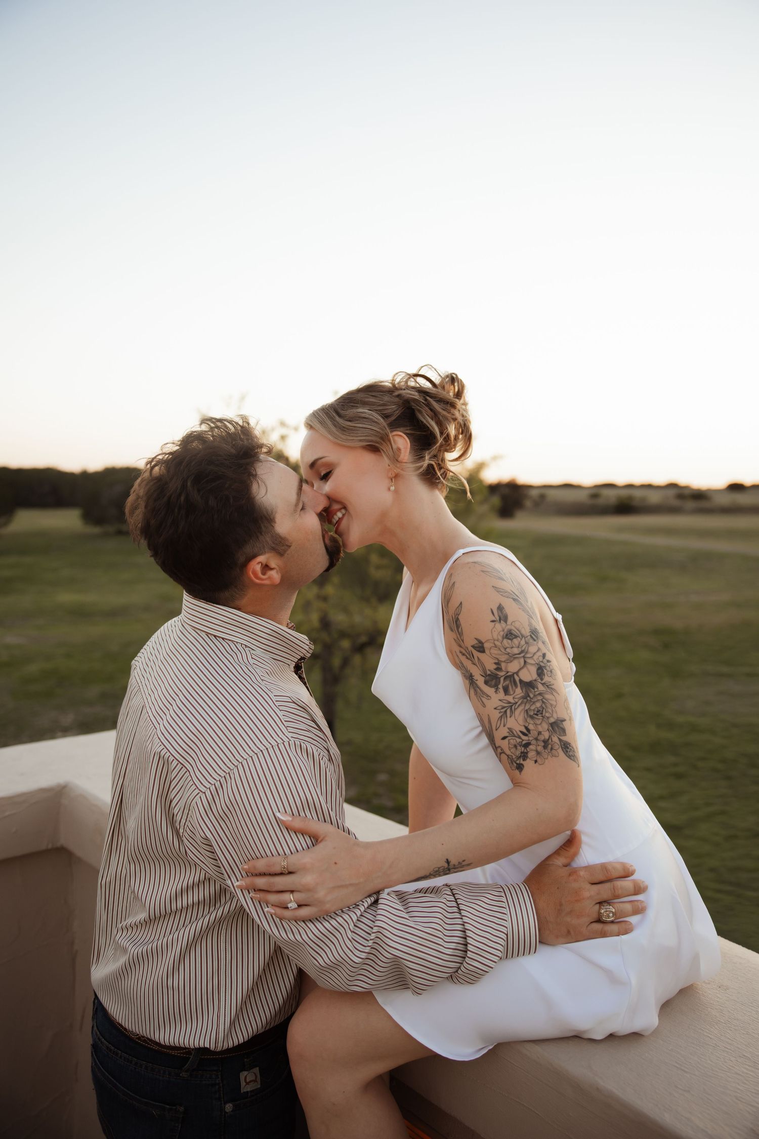Couple sharing a romantic kiss at sunset, woman in white dress with floral arm tattoo sitting on ledge embracing man in striped shirt.