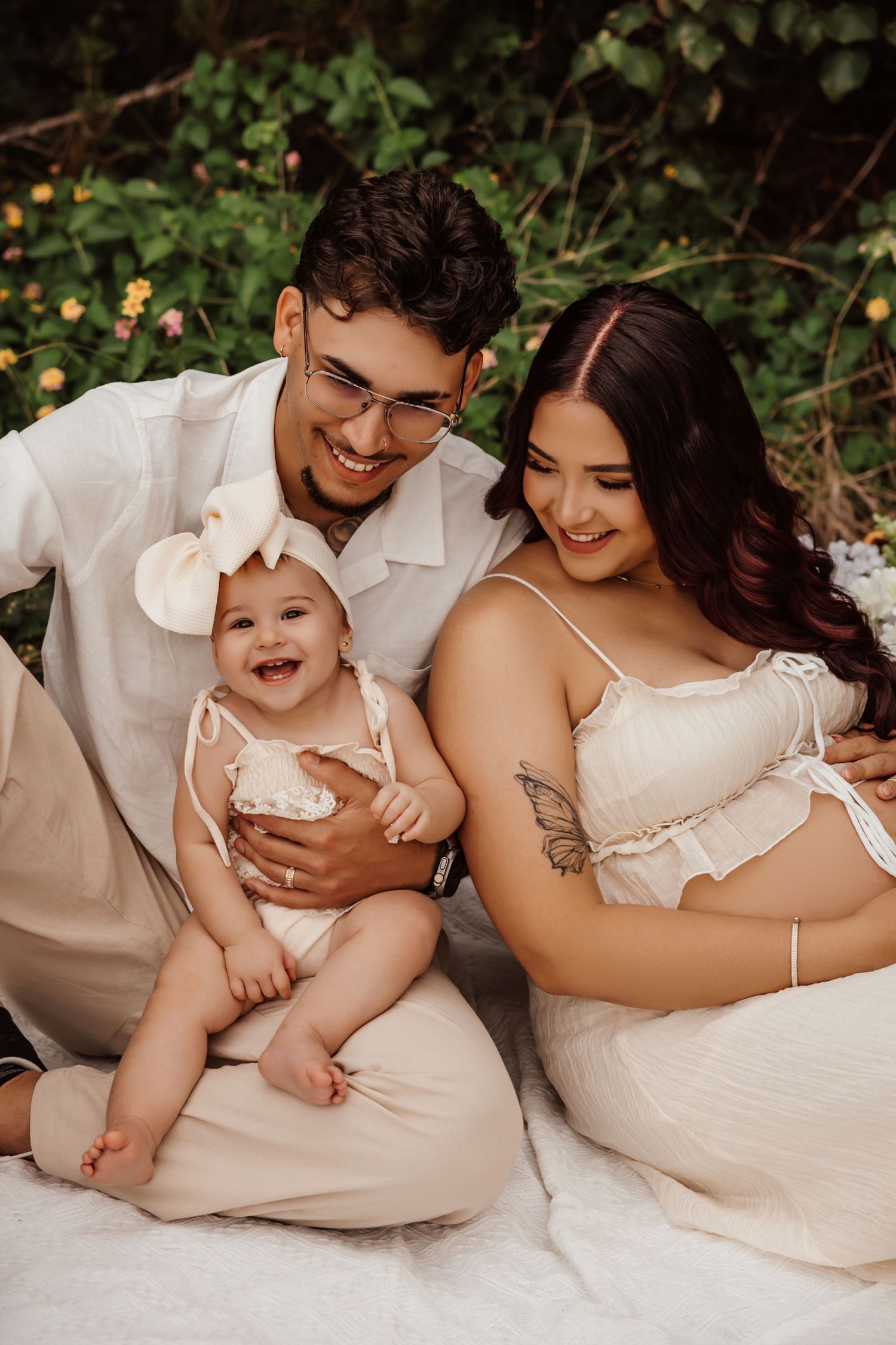 Young family with smiling baby girl in white bow sits outdoors among greenery for a beautiful maternity photo session.