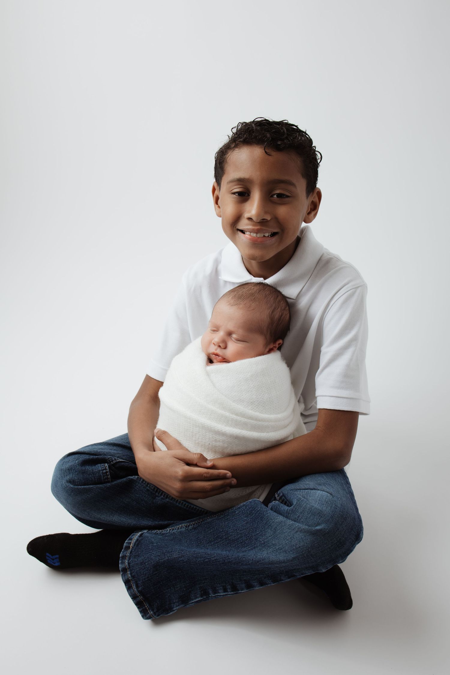 Smiling boy sitting cross-legged holding a swaddled newborn baby on a white background.