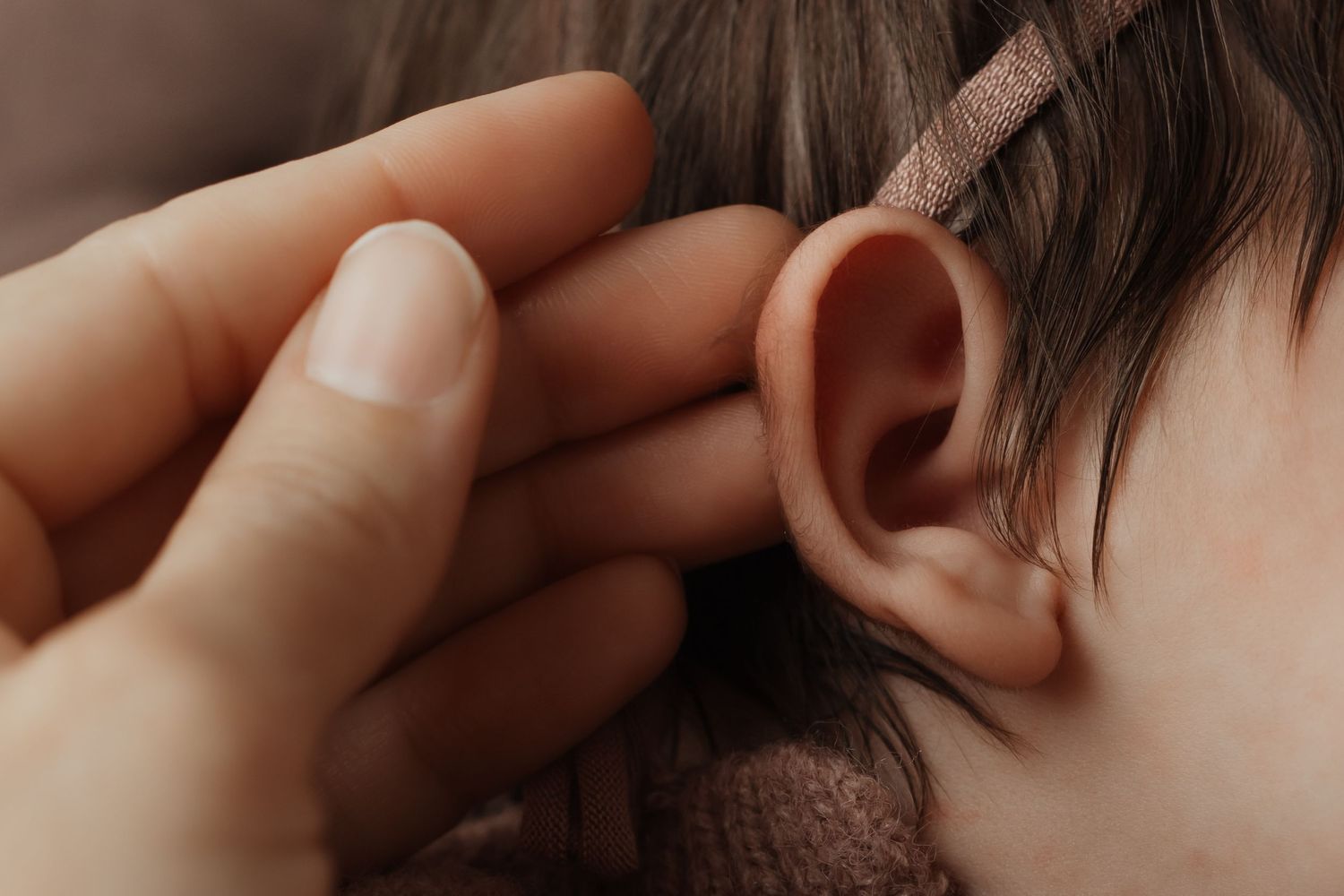 Adult hand gently touching a newborn babys tiny ear, with soft pink headband visible in dark hair.