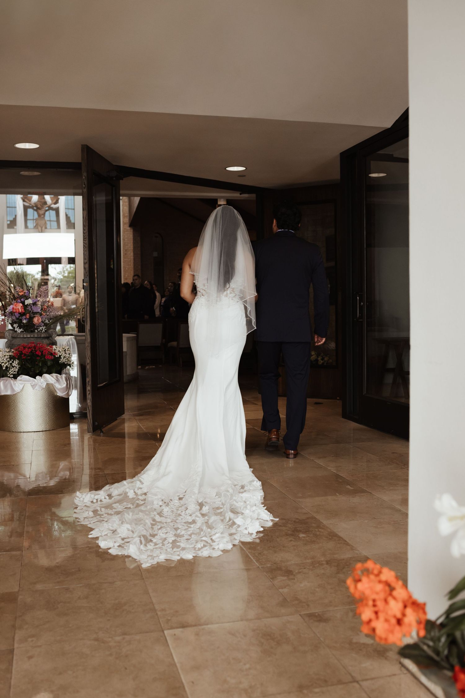 Bride in white mermaid gown with floral train and veil walks with groom in navy suit through elegant venue doors.