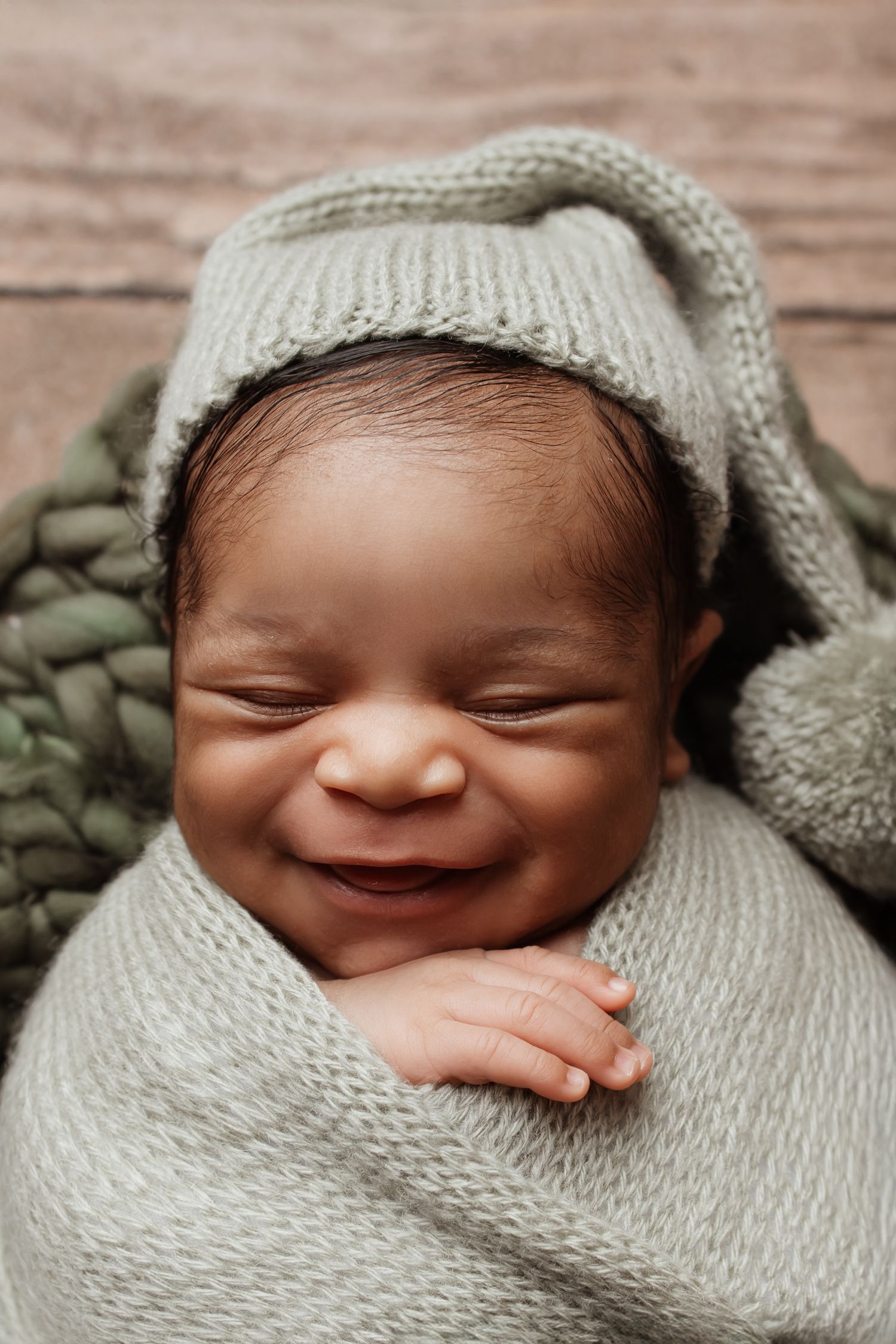 An adorable smiling newborn baby wrapped in a gray knit swaddle and matching sleepy hat poses cutely.