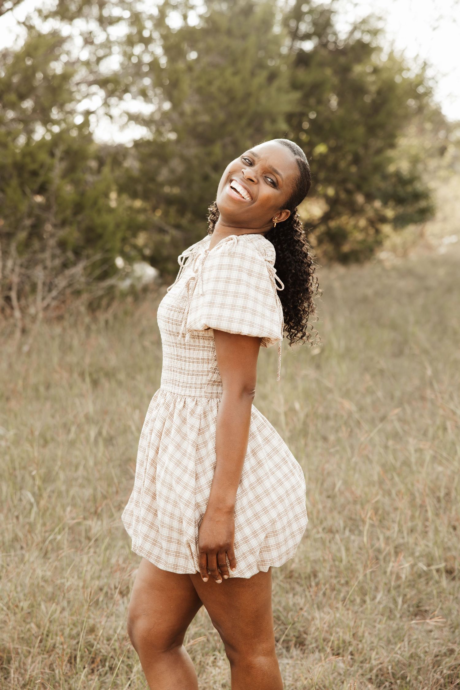 Young Black woman in plaid mini dress smiling outdoors in a grassy field.