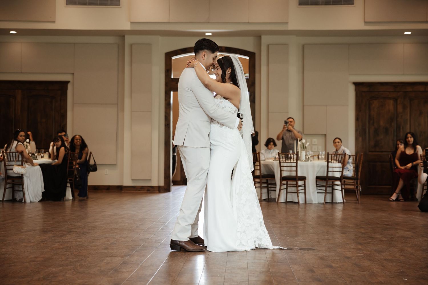 Bride and groom share their first dance in elegant ballroom with guests seated in background.
