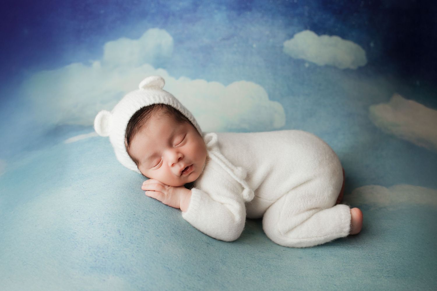 Sleeping newborn in white bear romper and bonnet on a dreamy blue sky backdrop.
