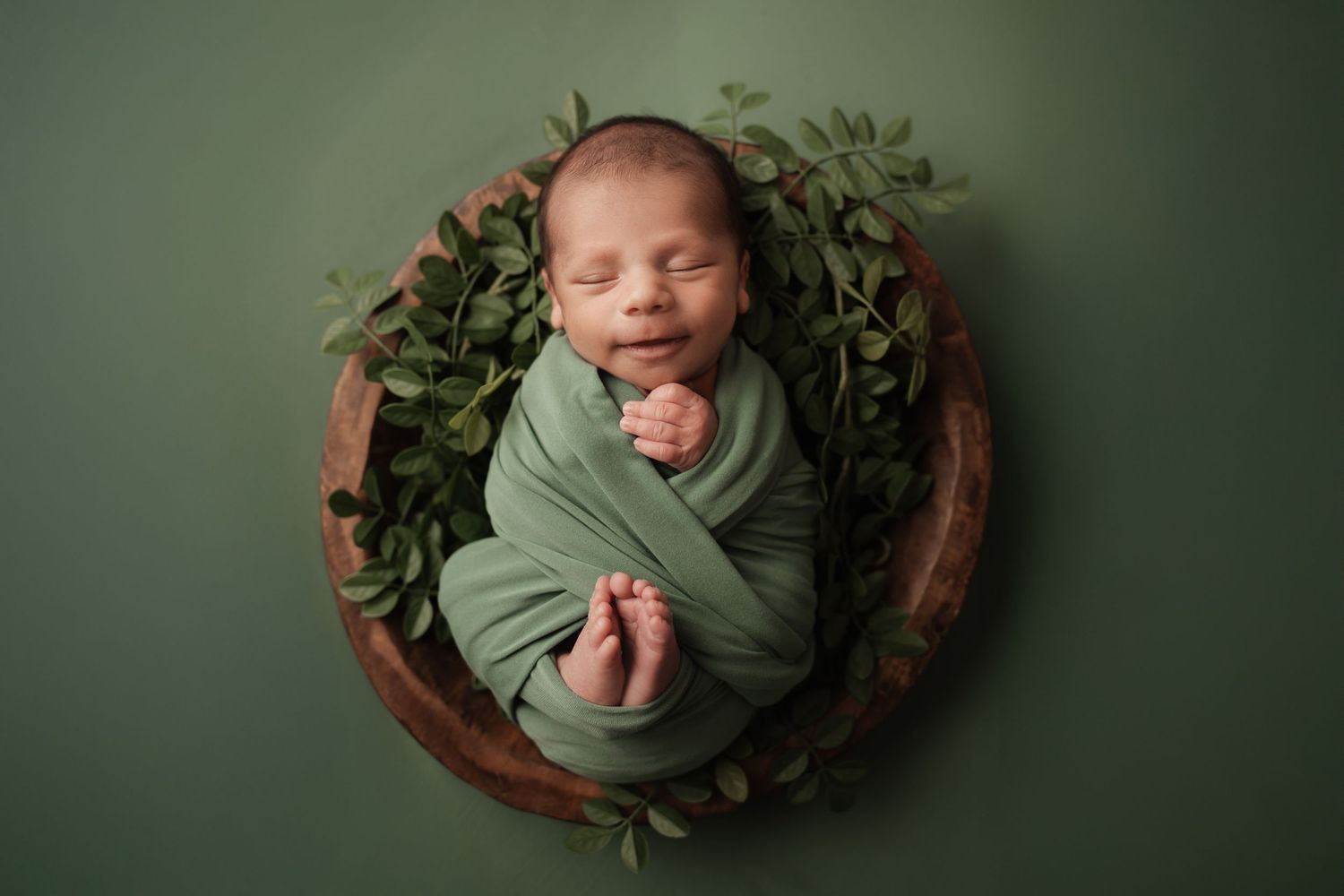 Smiling newborn baby swaddled in green wrap inside a wooden bowl with leafy greenery.