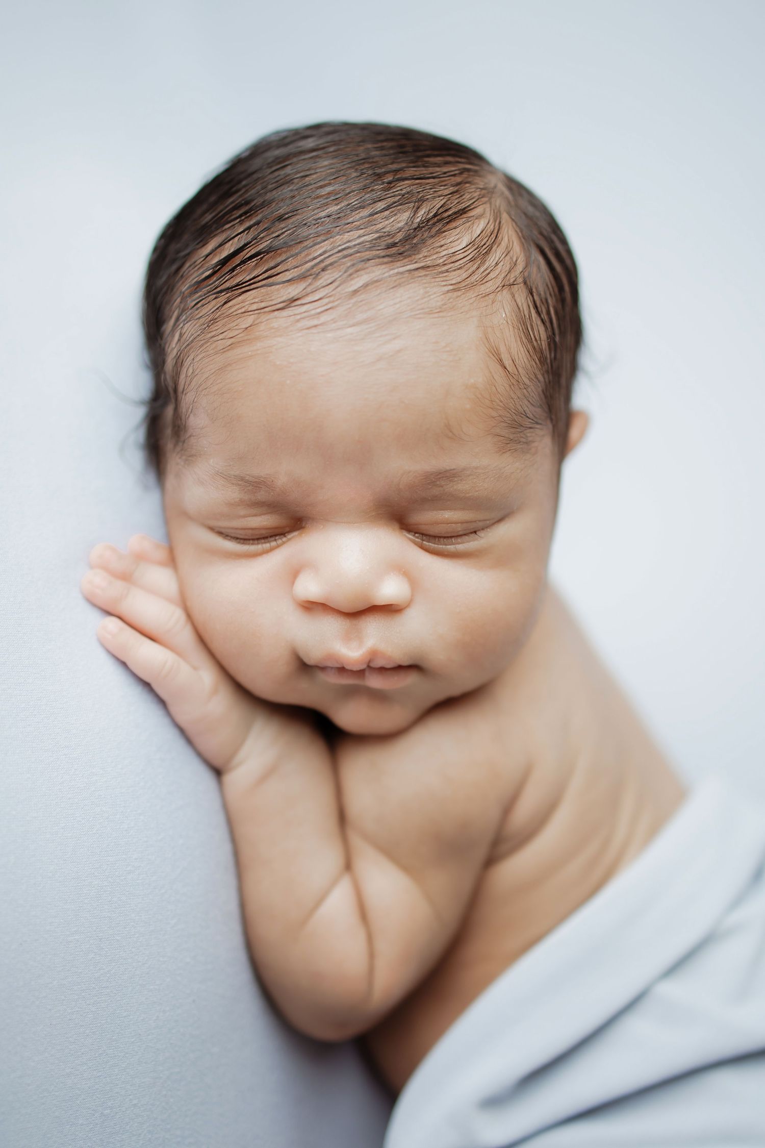 A peaceful newborn baby sleeps with hands tucked under chin on a soft light blue blanket.