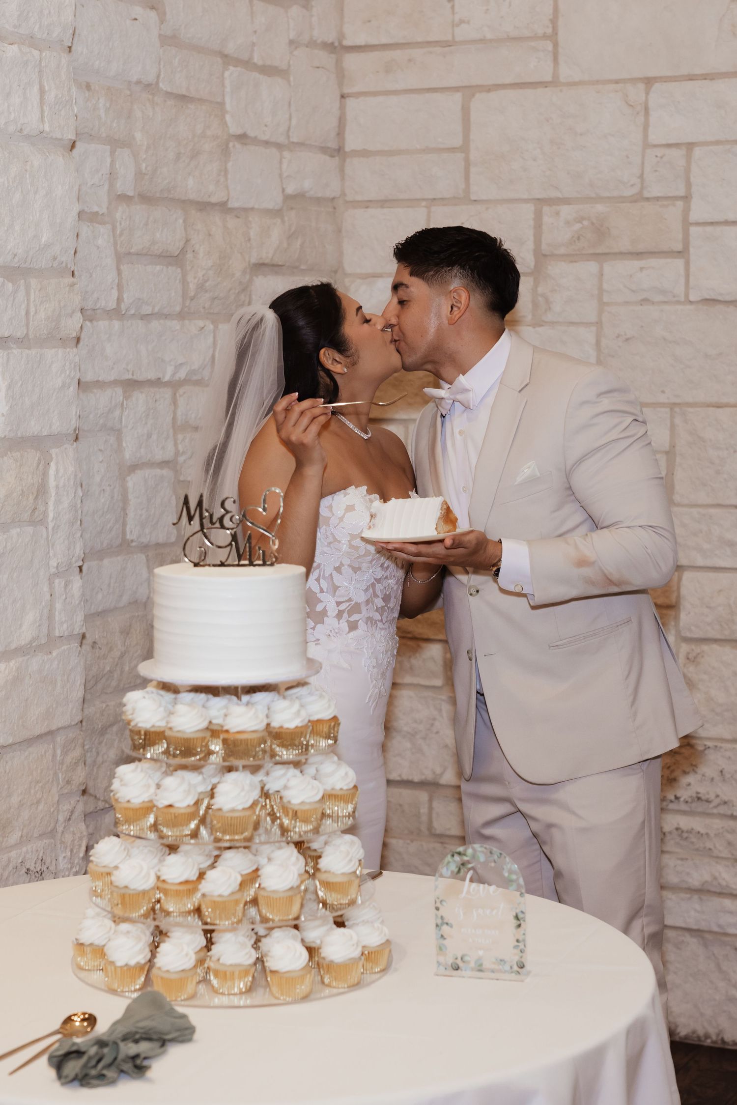 Bride and groom kissing behind tiered wedding cupcake display with white frosting and gold accents, Mr & Mrs cake topper.