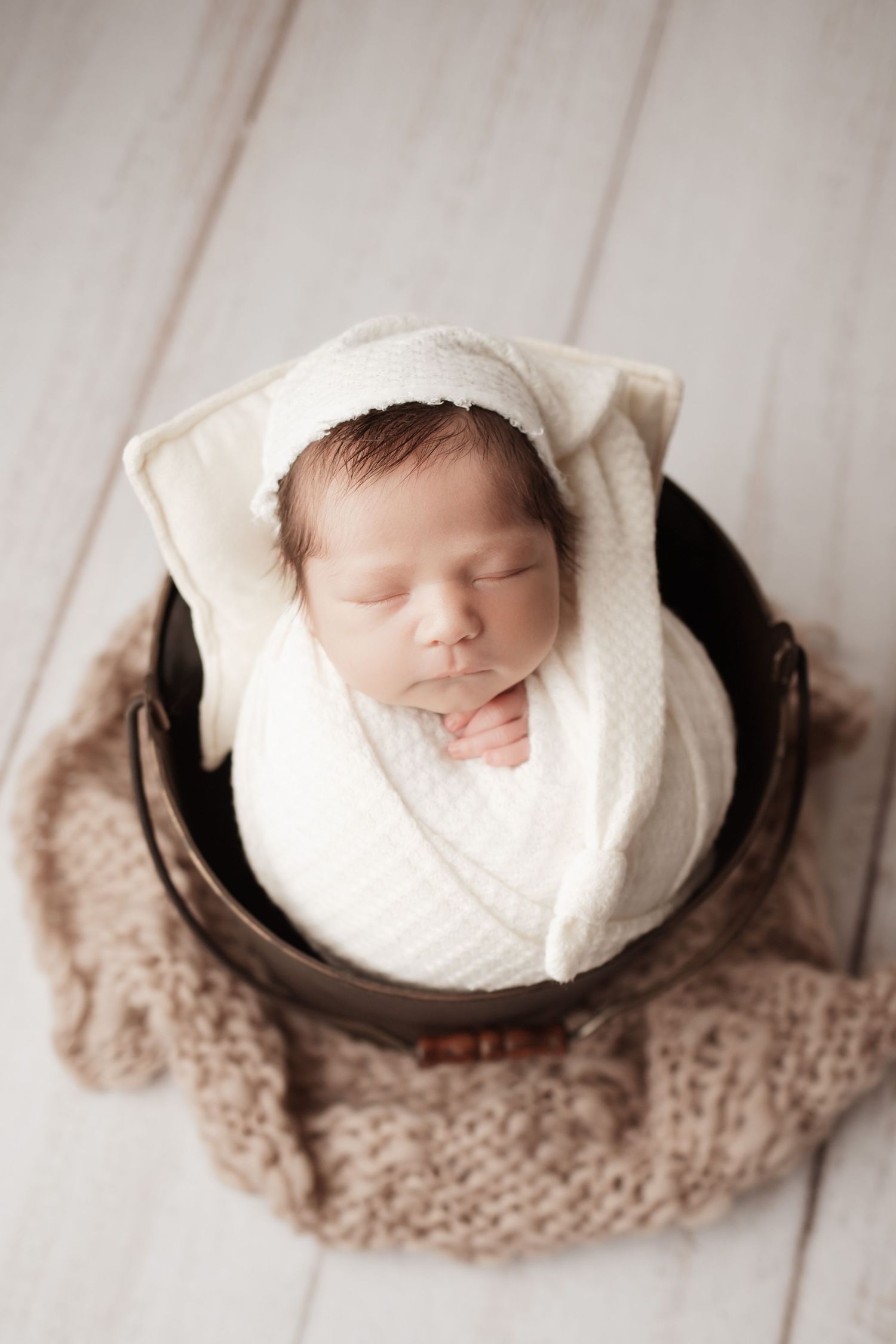 Sleeping newborn baby swaddled in white wrap with bonnet hat posed in wooden bucket prop.
