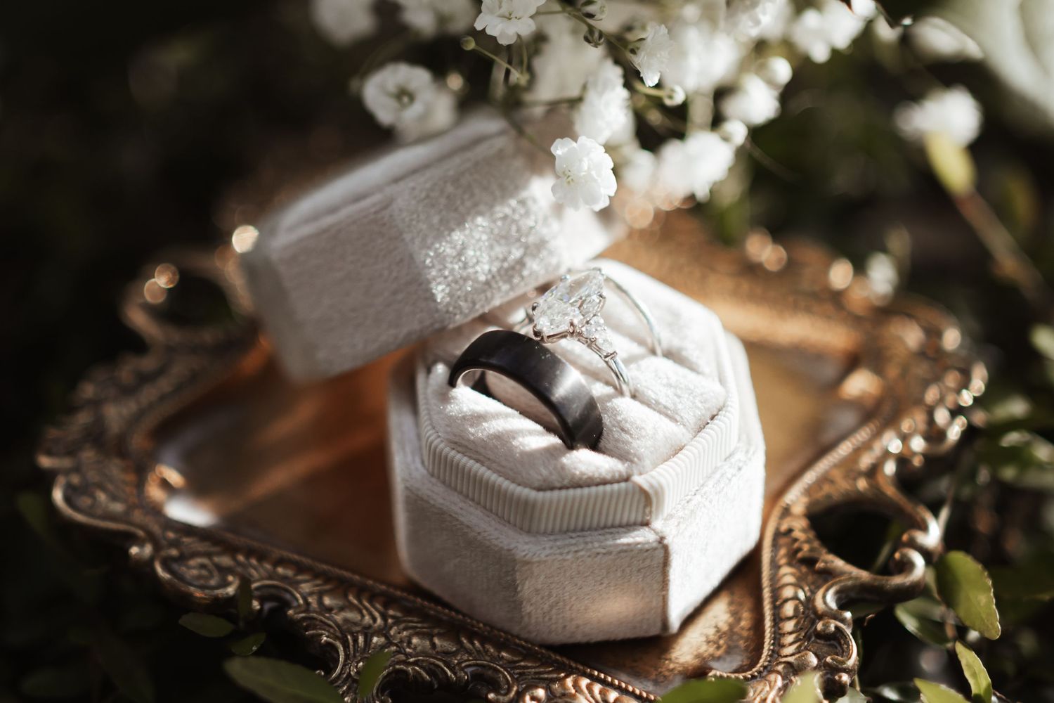A silver wedding band displayed in an elegant white velvet ring box on a ornate gold tray with white flowers.