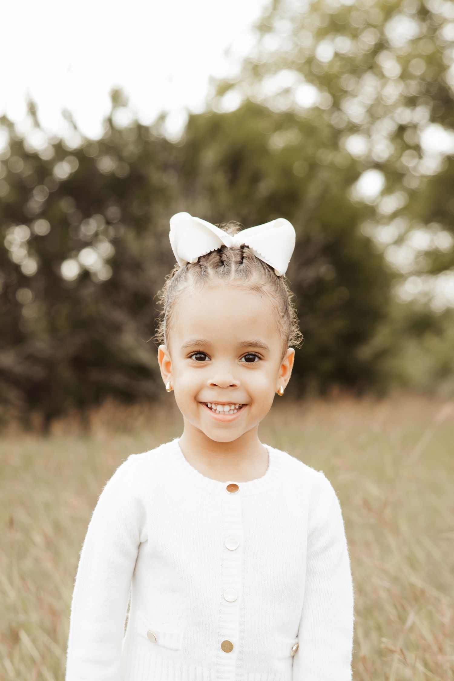 Smiling little girl with white bow in hair wearing white cardigan outdoors in a field.