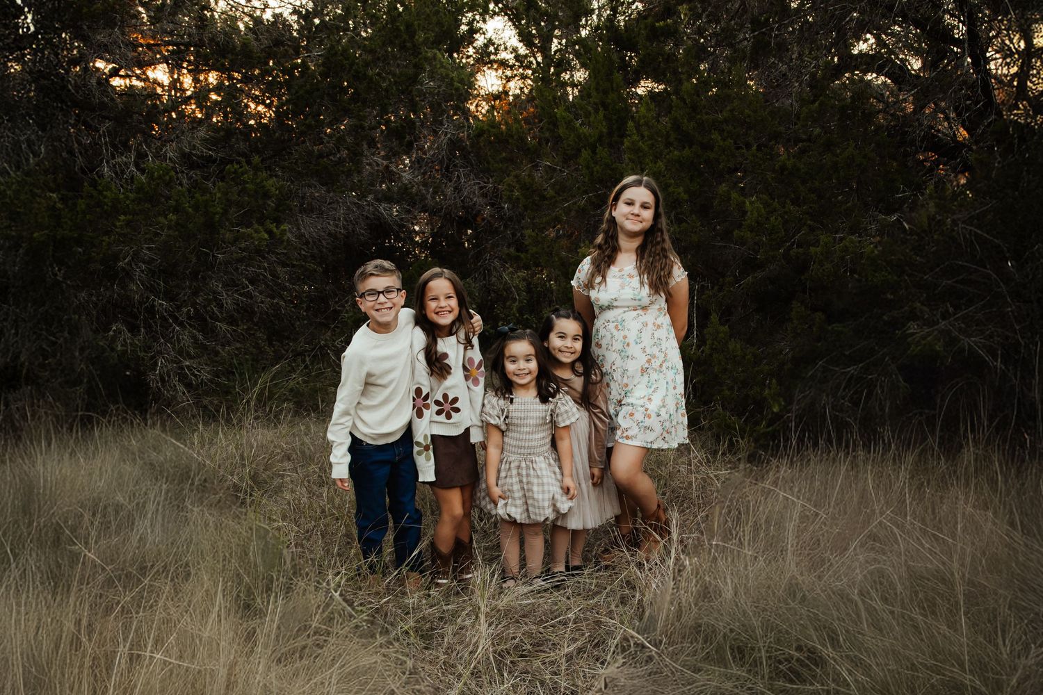 Five children pose together in a golden field surrounded by trees during a warm sunset outdoor photo session.