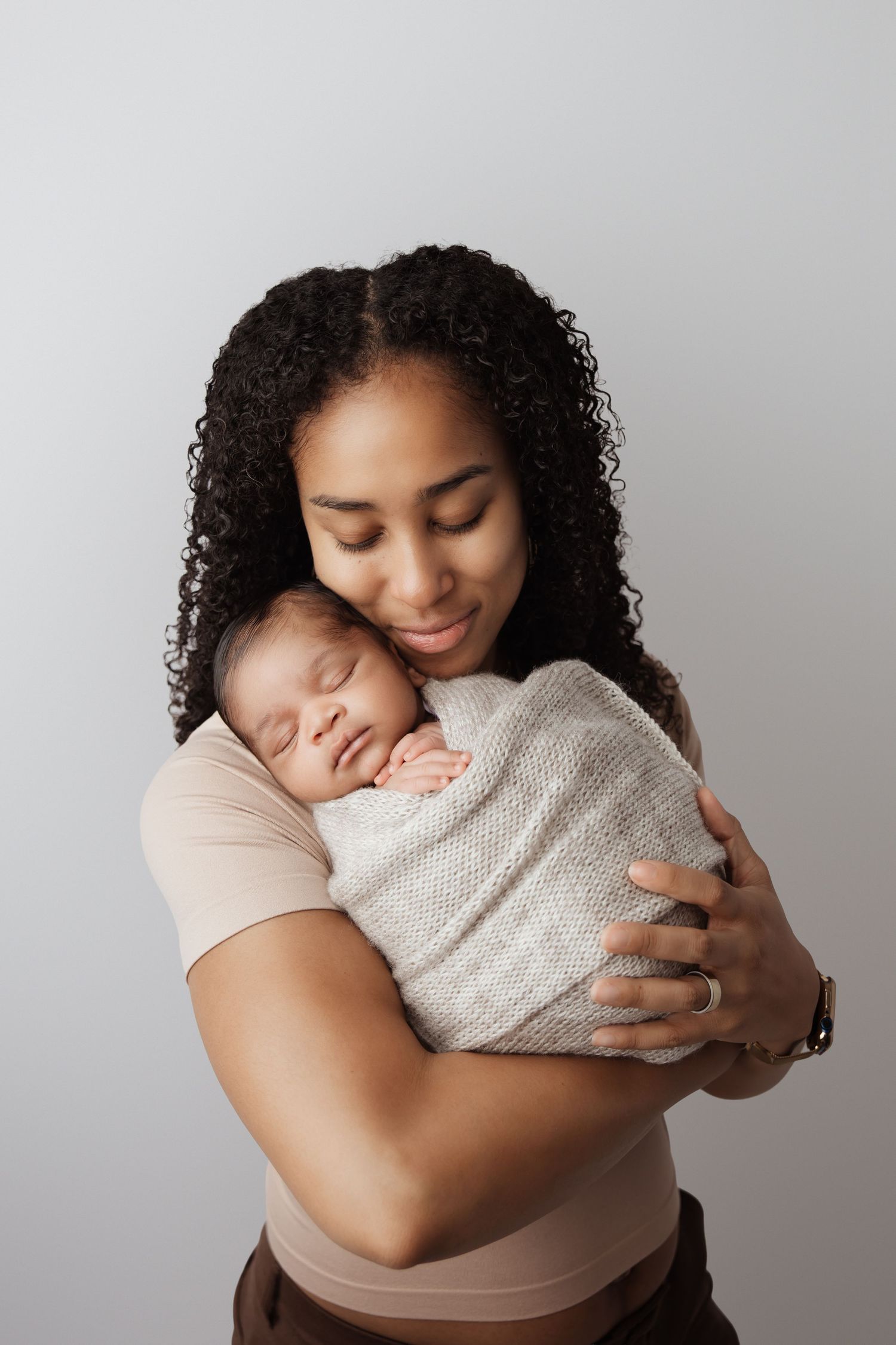 A mother with curly hair tenderly holds her swaddled newborn baby close to her cheek, smiling warmly.