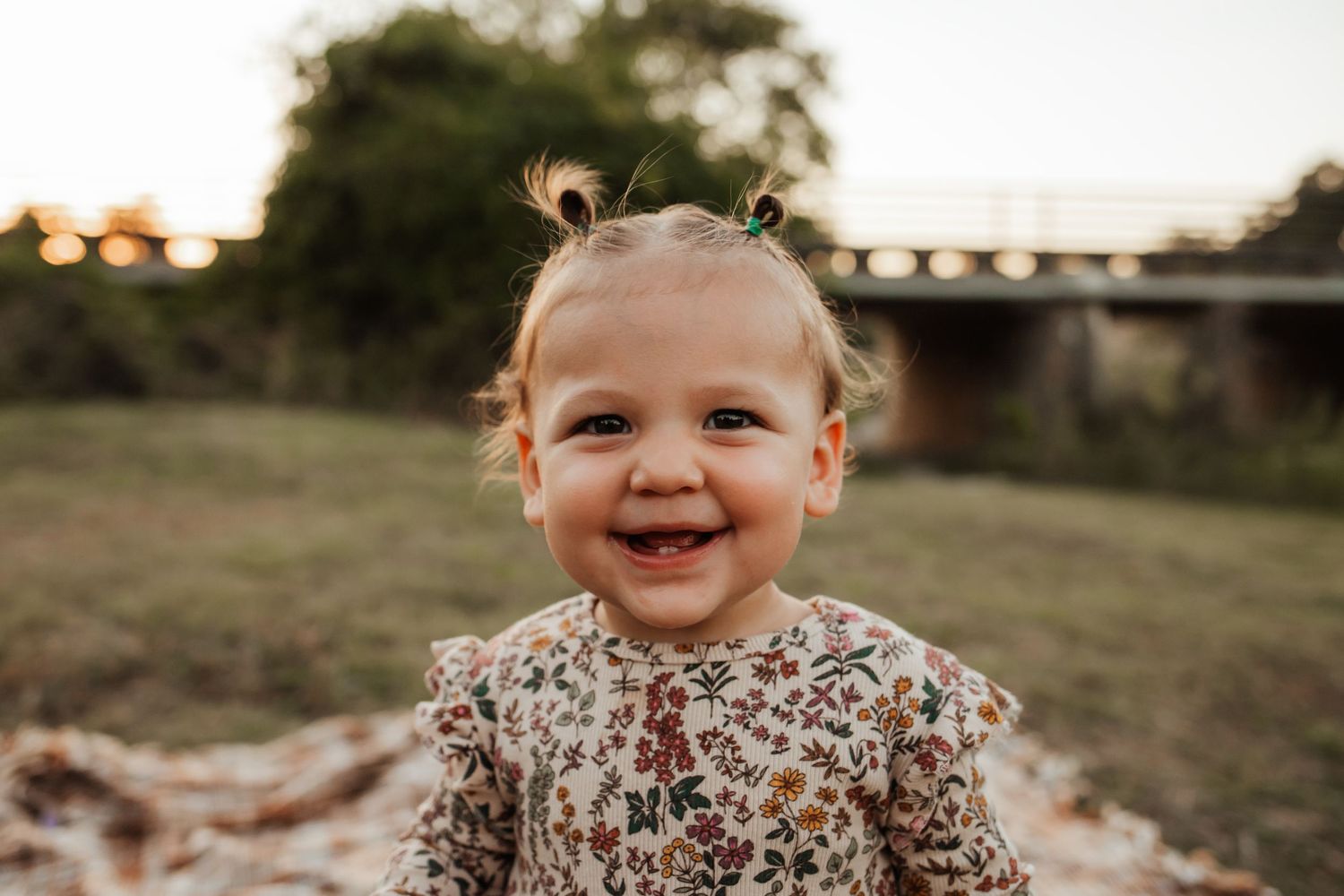 An adorable smiling baby girl with tiny pigtails sits outdoors during a golden hour portrait session.