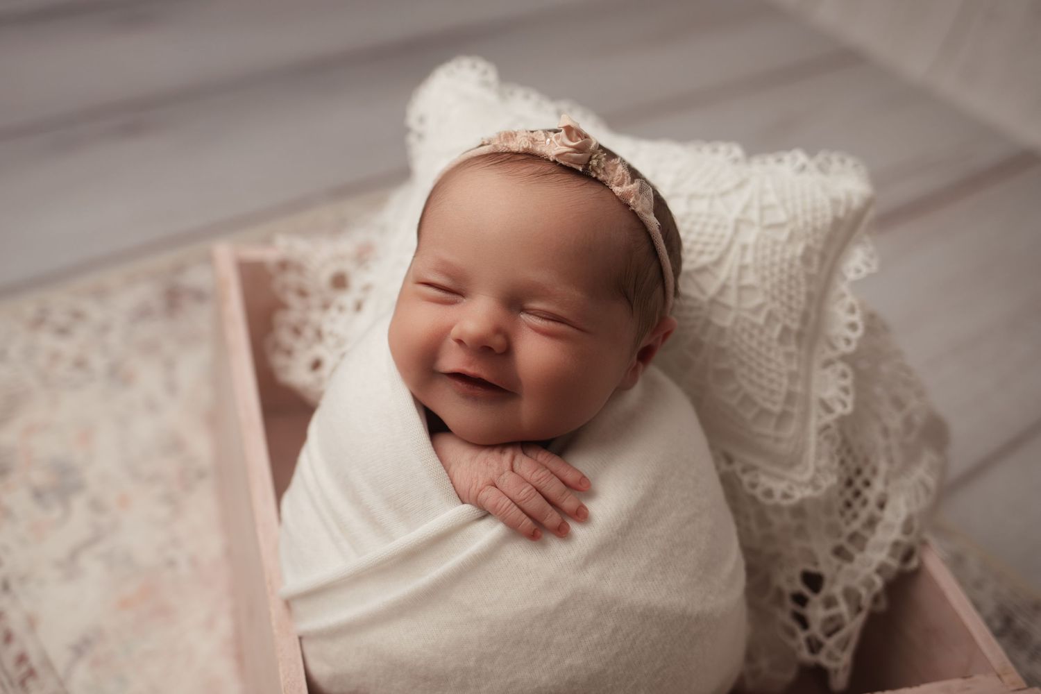Smiling newborn baby wrapped in white lace swaddle wearing a floral headband in a wooden box.