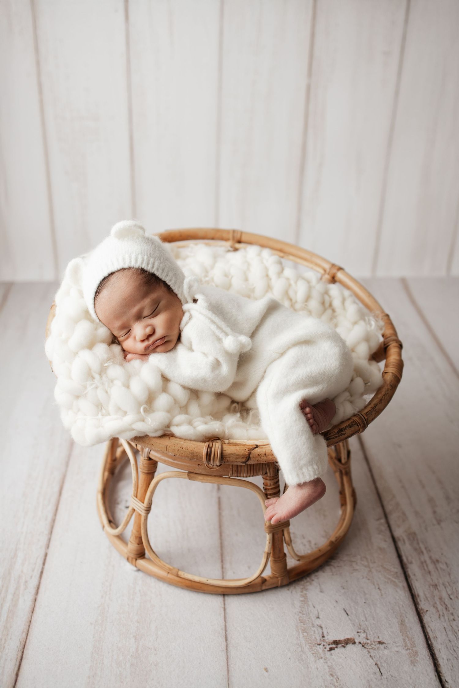 Newborn in white bear romper and bonnet sleeping in small round rattan chair with knit blanket.