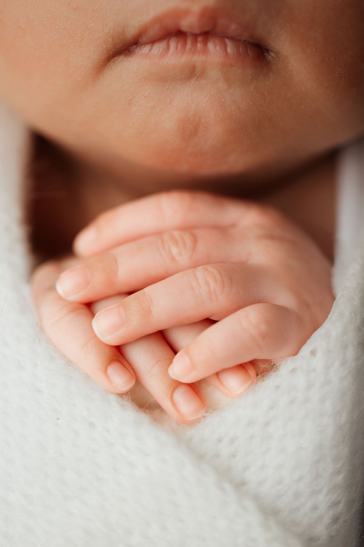Close-up of a sleeping newborn baby lips and tiny delicate hands wrapped in a soft white swaddle.