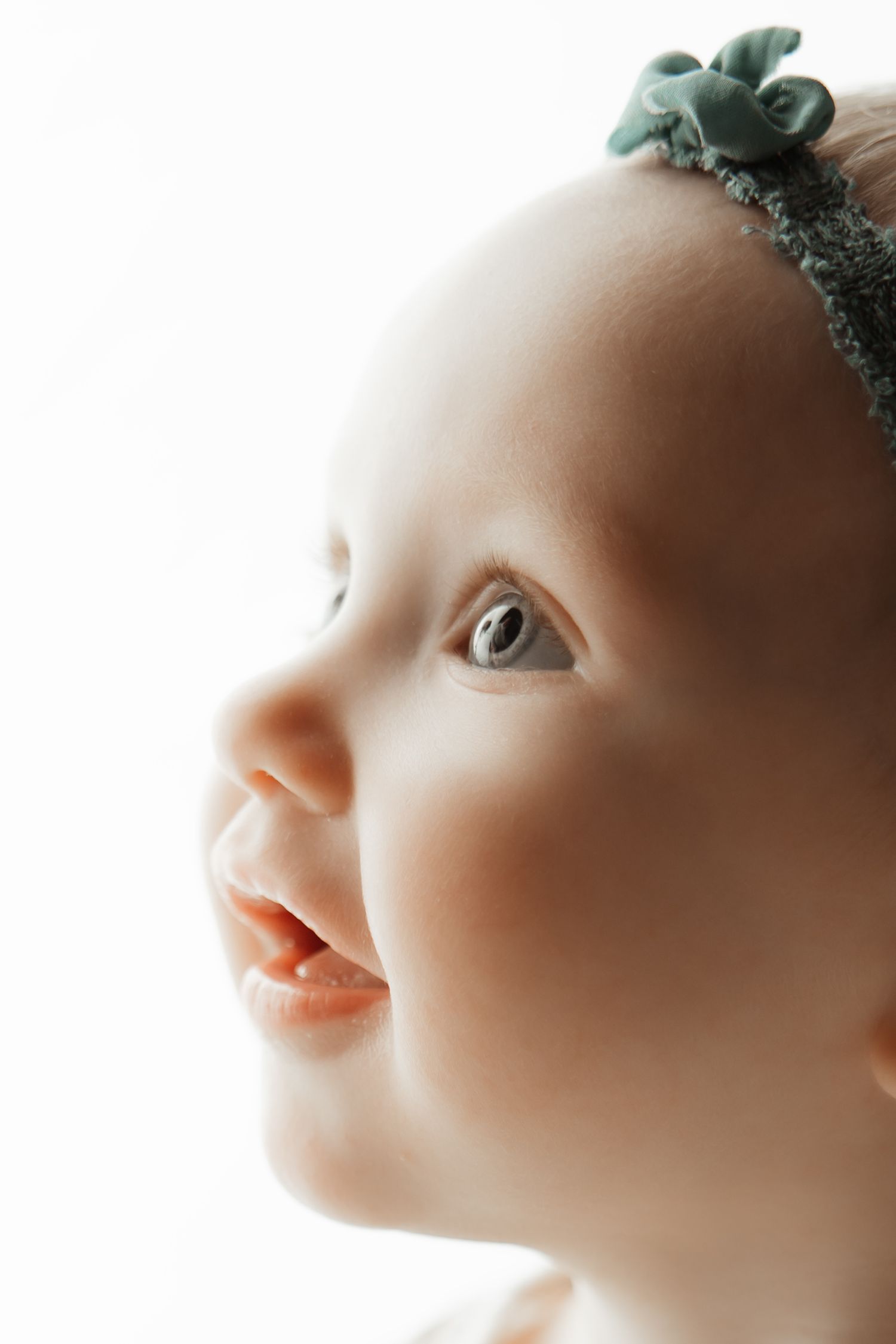 Close-up portrait of a smiling baby girl wearing a green floral headband, looking up with bright curious eyes.