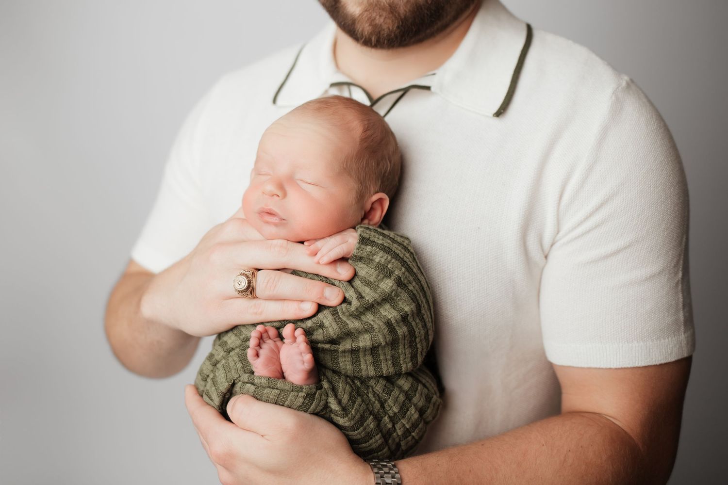 Father in white polo shirt cradling sleeping newborn baby wrapped in green striped blanket.
