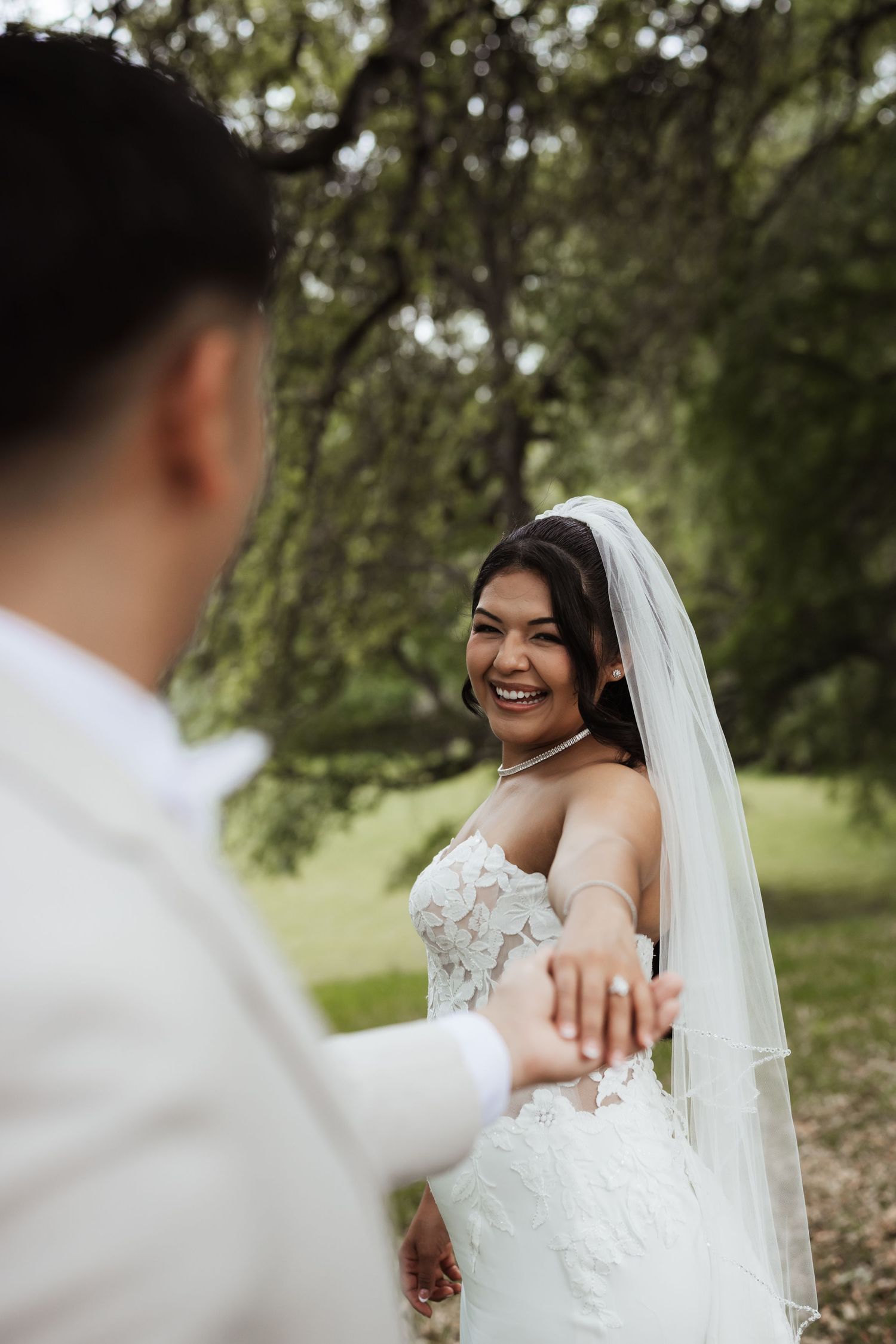 Smiling bride in lace gown and veil holds groom's hand outdoors among lush green trees on wedding day.