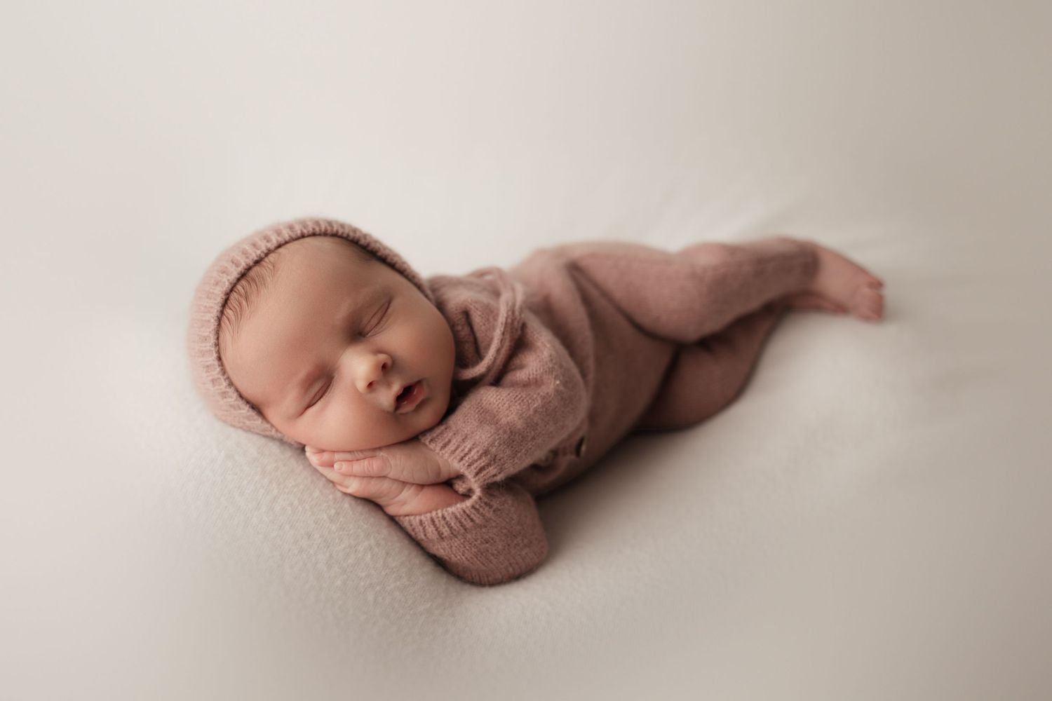 Sleeping newborn baby in pink knit outfit and bonnet posed on soft white blanket.