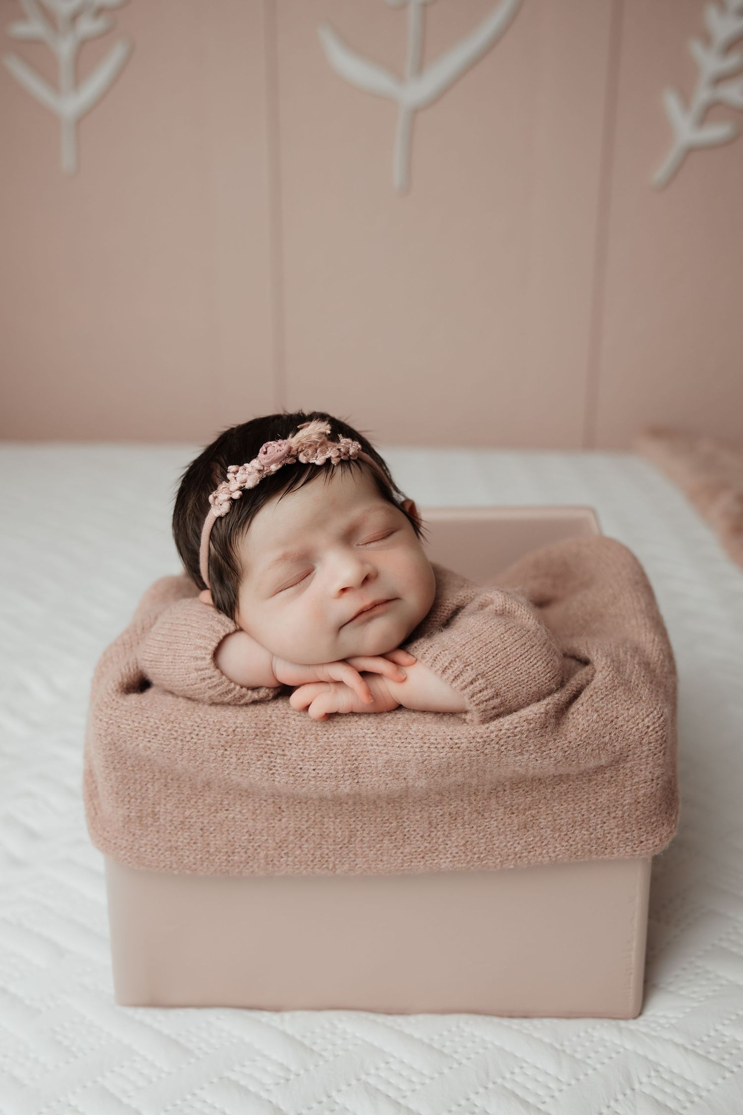 Newborn baby girl sleeping in a pink box wearing a floral headband and beige knit outfit.