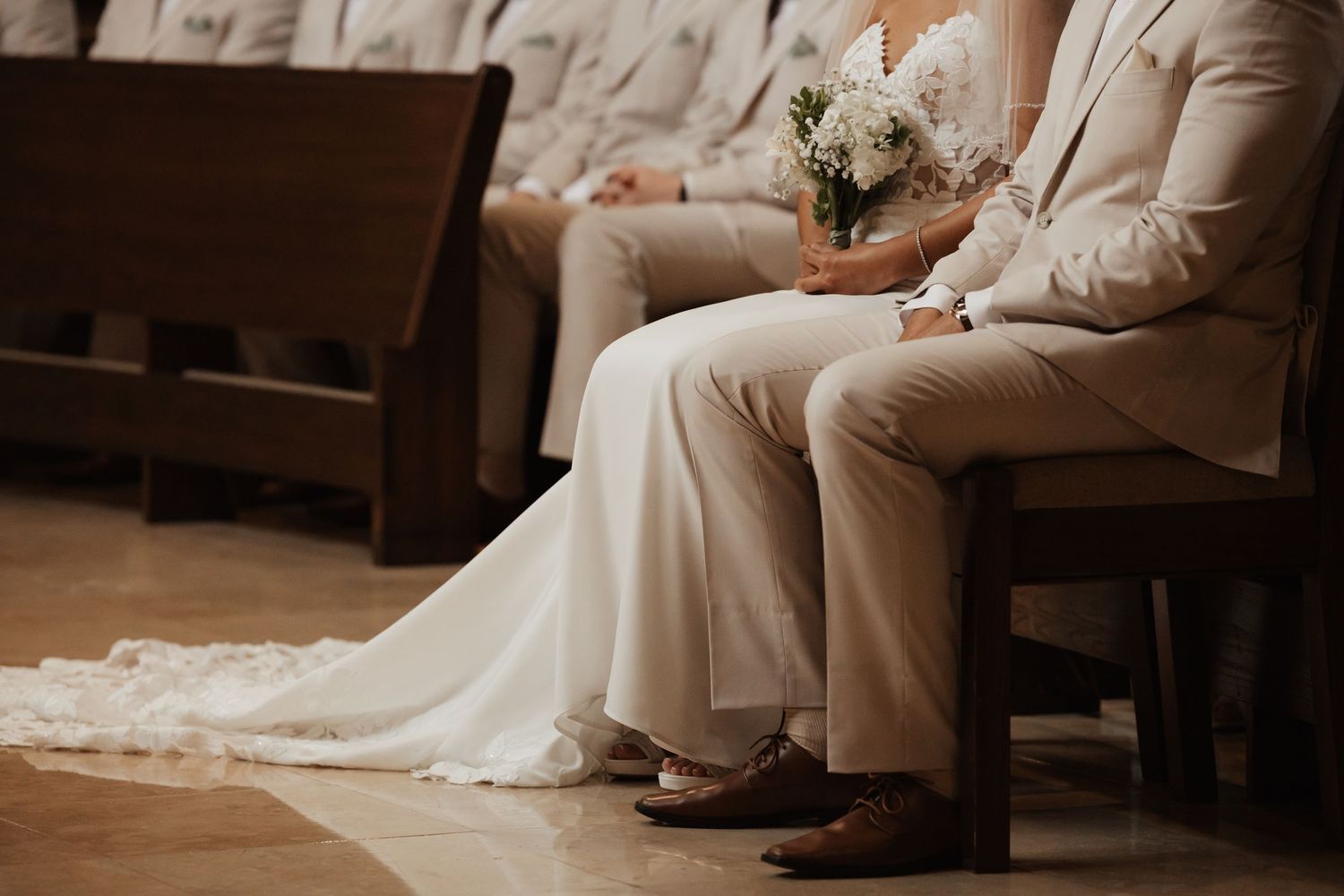Bride and groom sitting on wooden church pew, bride holding white bouquet, wearing elegant white gown and beige suit.