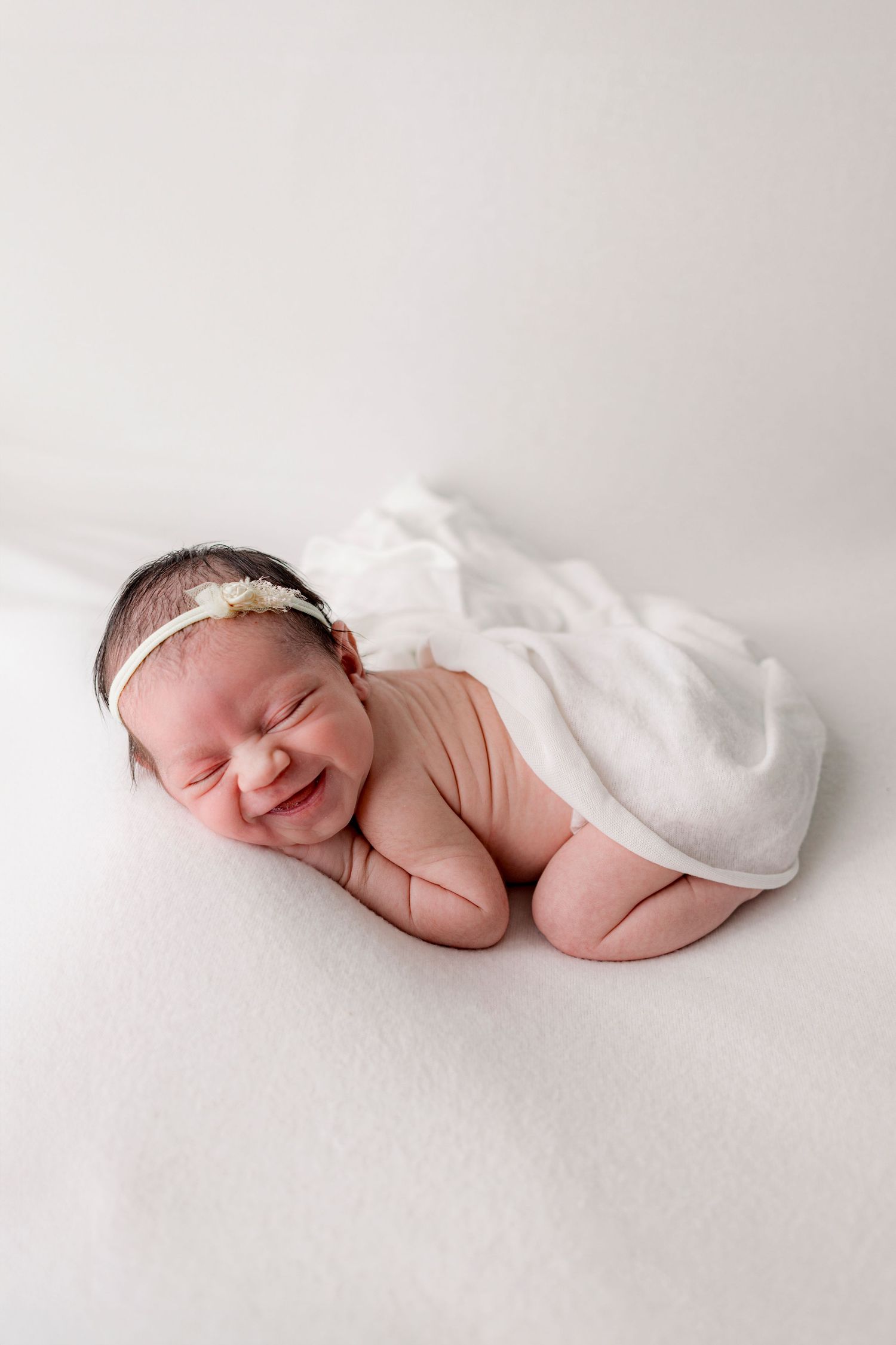 Smiling newborn baby girl with floral headband wrapped in white on a white blanket.