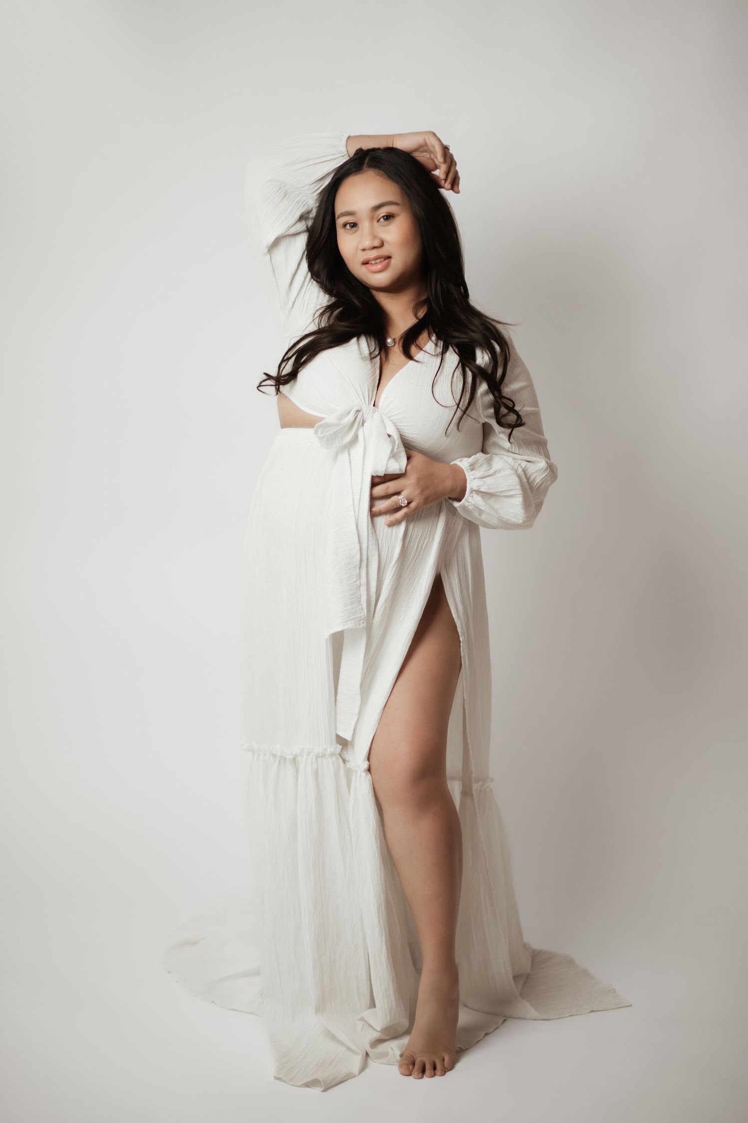 Pregnant woman in flowing white maternity gown posing gracefully against a white background.