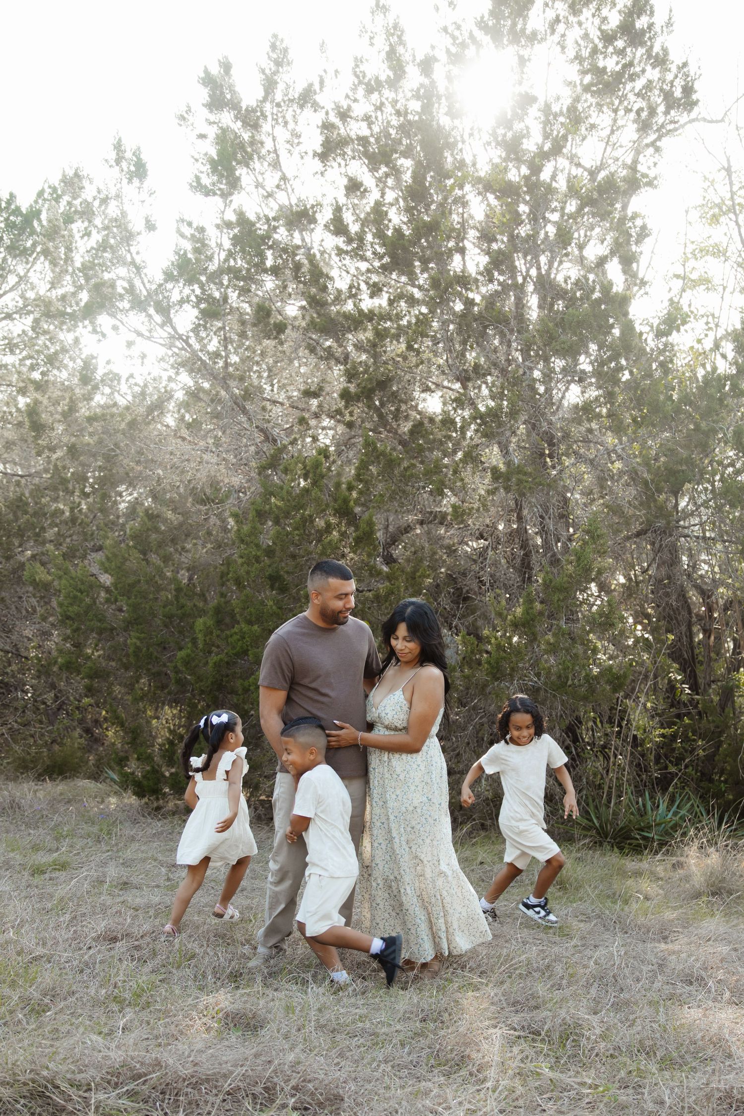 A family of five enjoys a sunny outdoor walk among trees, with parents and three young children dressed in white and neutral tones.