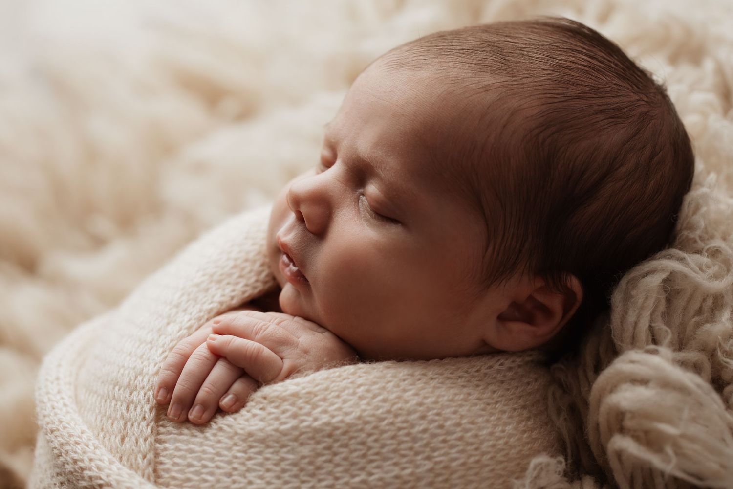 Close-up of a sleeping newborn baby wrapped in a cream knit blanket with tiny hands visible.