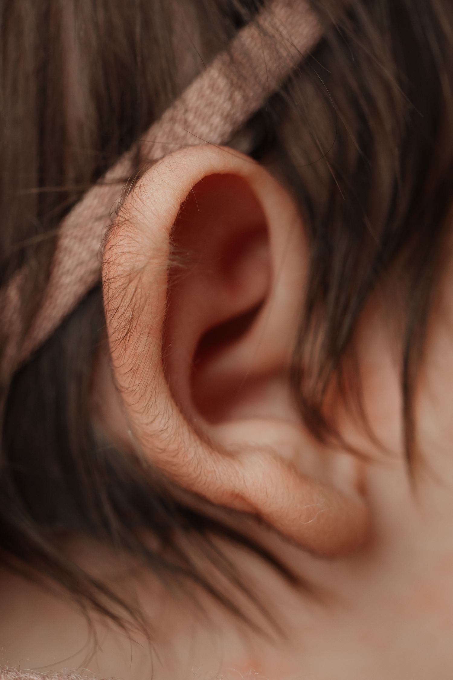 Detailed close-up of a newborn babys tiny ear with a soft pink headband visible in dark hair.